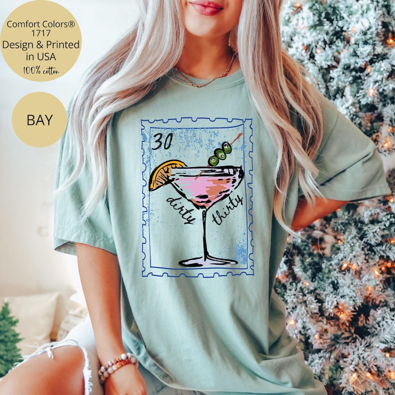May include: A light green t-shirt featuring a cocktail design with the text "30 dirty thirty". The design includes a pink cocktail with an olive garnish and a lemon slice. The shirt is made of 100% cotton.