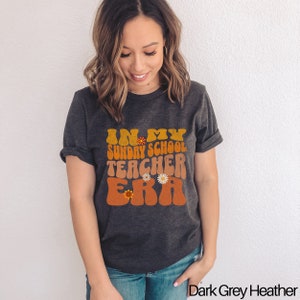 May include: A dark grey heather t-shirt with the text "In My Sunday School Teacher Era" in a retro font with orange and yellow accents and small flower graphics.
