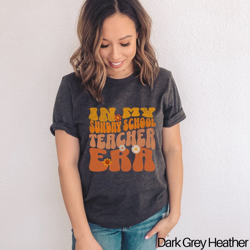 May include: A dark grey heather t-shirt with the text "In My Sunday School Teacher Era" in a retro font with orange and yellow accents and small flower graphics.