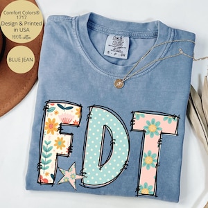 May include: Blue jean Comfort Colors t-shirt with the letters "FDT" in a floral and polka dot design. The shirt has a crew neck and short sleeves. The text "Comfort Colors" and "Design & Printed in USA" are also visible.
