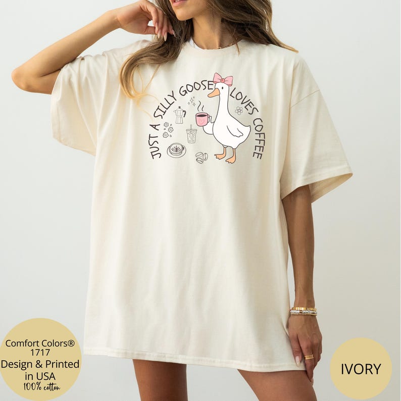 Puede incluir: Camiseta de color marfil con la frase "Just a Silly Goose Loves Coffee" y un ganso de dibujos animados con un lazo rosa, sosteniendo una taza de caf&eacute;. La camiseta es 100% algod&oacute;n e impresa en EE. UU.