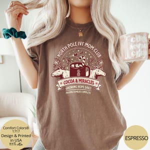 Puede incluir: Una camiseta marr&oacute;n con un dise&ntilde;o gr&aacute;fico de una taza de chocolate caliente con malvaviscos y un bast&oacute;n de caramelo. El texto en la camiseta dice "North Pole IVF Mom Club *Cocoa & Miracles* Growing Hope Daily Making Families Complete".