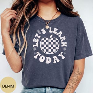May include: A denim-colored t-shirt with the text "LET'S LEARN TODAY" in a circular design. The design includes a checkered apple graphic in the center. The shirt is a comfortable, casual style.
