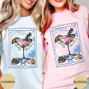 K&ouml;nnte beinhalten: Hellblaue und rosa T-Shirts mit einem "Dirty Martini"-Motiv im Briefmarken-Design. Die Illustration zeigt ein Martiniglas mit Oliven und einer Zitronenscheibe. Die Shirts sind mit "Chambray" und "Blossom" beschriftet.