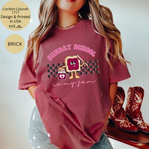 May include: A maroon t-shirt with a graphic of a slice of bread with a heart-shaped jar of jam and the text "Sunday School is my jam".