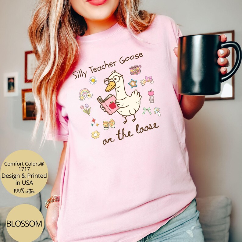 Pode incluir: Camiseta rosa com um ganso de desenho animado usando &oacute;culos e segurando um livro. O texto "Silly Teacher Goose on the loose" est&aacute; impresso na camiseta.