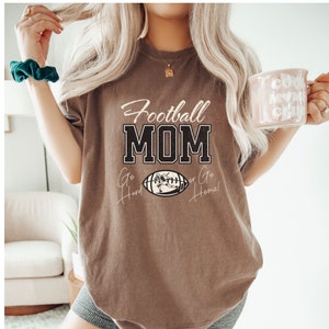 May include: A brown t-shirt with the text "Football MOM" in black letters. The shirt also features a football graphic with the text "Go Hard or Go Home!" in a cursive font.