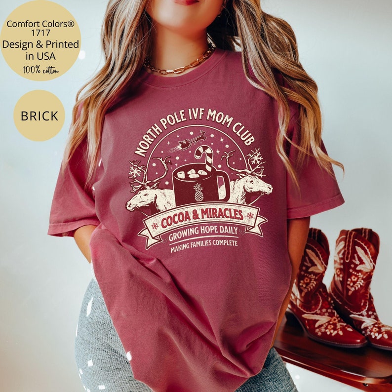 Puede incluir: Una camiseta roja con un dise&ntilde;o gr&aacute;fico blanco que dice "North Pole IVF Mom Club" con una taza de chocolate caliente y renos. El texto debajo del gr&aacute;fico dice "Cocoa & Miracles", "Growing Hope Daily", y "Making Families Complete".