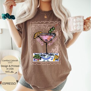 K&ouml;nnte beinhalten: Espressofarbenes T-Shirt mit einem Grafikdesign eines Dirty Martini-Cocktails, Oliven und einer Zitronenscheibe, eingerahmt in einem Poststempel-&auml;hnlichen Rahmen. Hergestellt aus 100% Baumwolle.