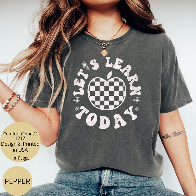 May include: Pepper-colored t-shirt with the text "LET'S LEARN TODAY" in white, surrounding a checkered apple graphic. The shirt is made of 100% cotton and is printed in the USA. The shirt is a Comfort Colors 1717 design.