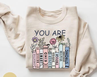 May include: Sand-colored sweatshirt with the words "YOU ARE" in navy blue. Below the text are illustrated books with positive words like "Creative," "Brave," and "Strong." Colorful flowers and foliage are also depicted.