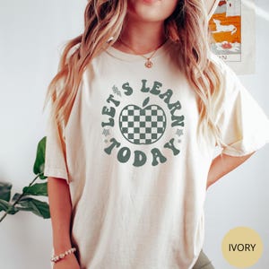 May include: Ivory-colored t-shirt with the text "Let's Learn Today" in a circular design. The design includes an apple with a checkered pattern. The shirt is a relaxed fit.