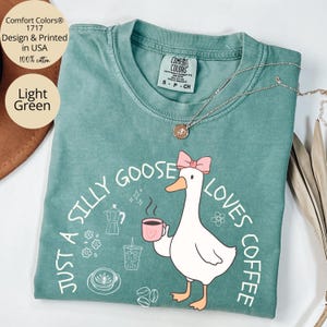 Puede incluir: Camiseta Comfort Colors verde claro con el texto "Just a Silly Goose Loves Coffee" y un dibujo de un ganso de dibujos animados sosteniendo una taza de caf&eacute;. La camiseta es 100% algod&oacute;n y est&aacute; dise&ntilde;ada e impresa en EE. UU.