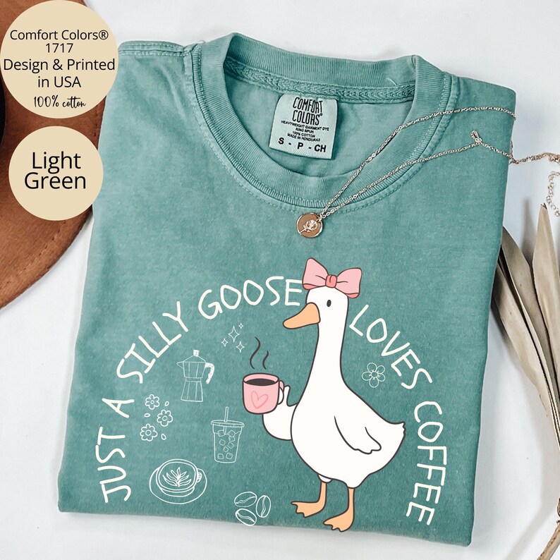 Puede incluir: Camiseta Comfort Colors verde claro con el texto "Just a Silly Goose Loves Coffee" y un dibujo de un ganso de dibujos animados sosteniendo una taza de caf&eacute;. La camiseta es 100% algod&oacute;n y est&aacute; dise&ntilde;ada e impresa en EE. UU.