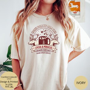 Puede incluir: Una camiseta de color crema con un dise&ntilde;o gr&aacute;fico rojo y blanco que dice "North Pole IVF Mom Club" con una taza de chocolate caliente y renos. El texto debajo del gr&aacute;fico dice "Cocoa & Miracles", "Growing Hope Daily", y "Making Families Complete".