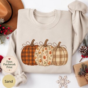 May include: A sand-colored crewneck sweatshirt featuring a central graphic of three pumpkins with a faux patchwork print. The pumpkins display orange plaid, autumn leaf patterns, and tan gingham designs with visible stitched outlines.