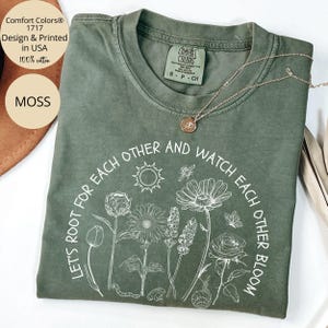 May include: A green t-shirt with a white graphic print of flowers and the text "Let's root for each other and watch each other bloom".