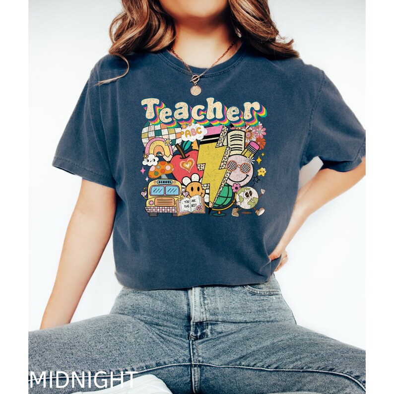May include: A navy blue t-shirt with the word "Teacher" in rainbow letters and a colorful graphic of school supplies, including a school bus, an apple, a lightning bolt, a book, a pencil, and a globe.