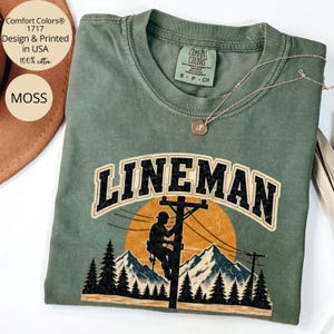 May include: A moss green crewneck t-shirt featuring a graphic of a lineman working on a utility pole against a large orange sun and mountain landscape. The text LINEMAN is printed in a bold, distressed black font above the graphic illustration.