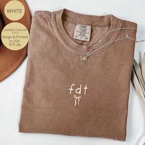 Peut inclure: Un t-shirt marron Comfort Colors avec les lettres "fdt" et un motif de n&oelig;ud brod&eacute; en blanc. Un collier argent&eacute; avec un pendentif repose sur le t-shirt. Le t-shirt est &eacute;tiquet&eacute; "Design & Printed in USA 100% cotton."