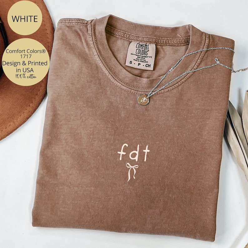 Peut inclure: Un t-shirt marron Comfort Colors avec les lettres "fdt" et un motif de n&oelig;ud brod&eacute; en blanc. Un collier argent&eacute; avec un pendentif repose sur le t-shirt. Le t-shirt est &eacute;tiquet&eacute; "Design & Printed in USA 100% cotton."