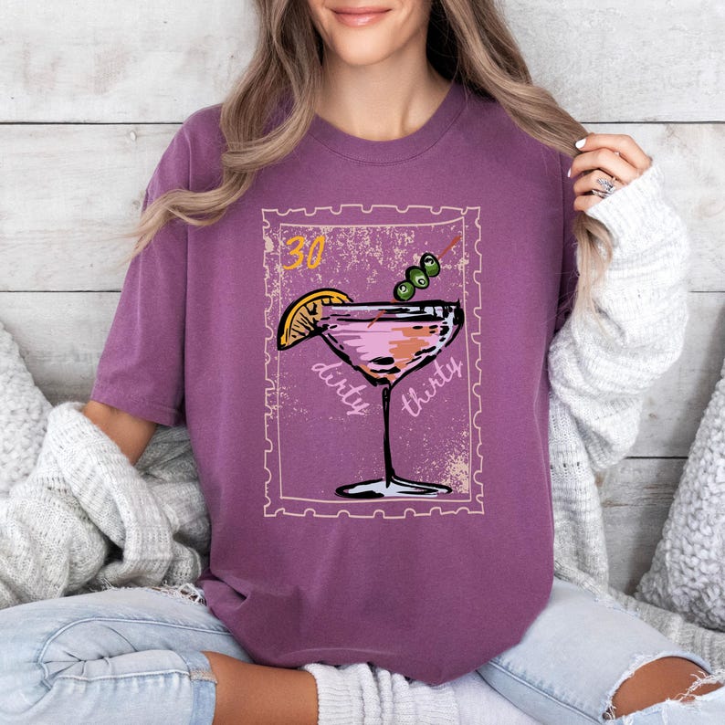 May include: A purple t-shirt featuring a cocktail design with the words "dirty thirty" and the number "30". The cocktail has a lemon slice and olives. The design is in a postage stamp style.
