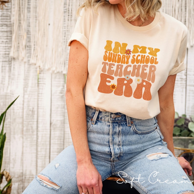 May include: A beige t-shirt with a retro-style graphic that says "In My Sunday School Teacher Era" with orange and yellow flowers.