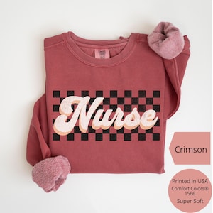 K&ouml;nnte beinhalten: Karmesinrotes Sweatshirt mit dem Wort "Nurse" in einem Retro-Schriftzug &uuml;ber einem schwarz-wei&szlig;en Schachbrettmuster. Das Sweatshirt besteht aus einem weichen Material und hat einen Rundhalsausschnitt. Der Text "Printed in USA Comfort Colors&reg; 1566 Super Soft" ist ebenfalls sichtbar.