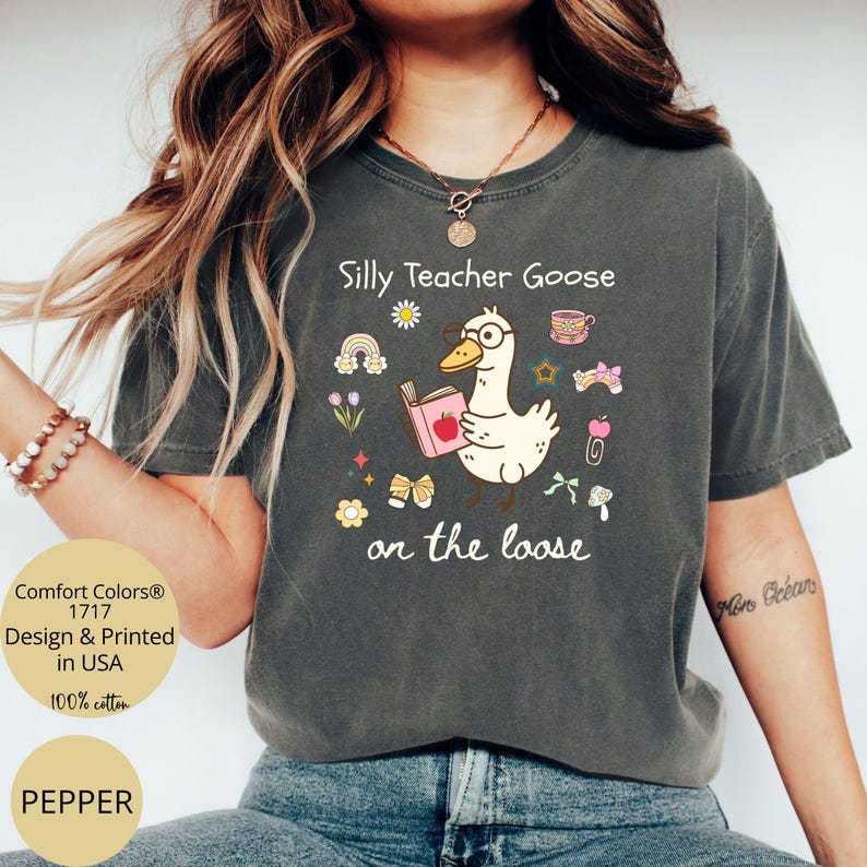 Pode incluir: Uma camiseta cinza escura com um ganso de desenho animado usando &oacute;culos e segurando um livro. O texto "Silly Teacher Goose on the loose" est&aacute; impresso na camiseta.