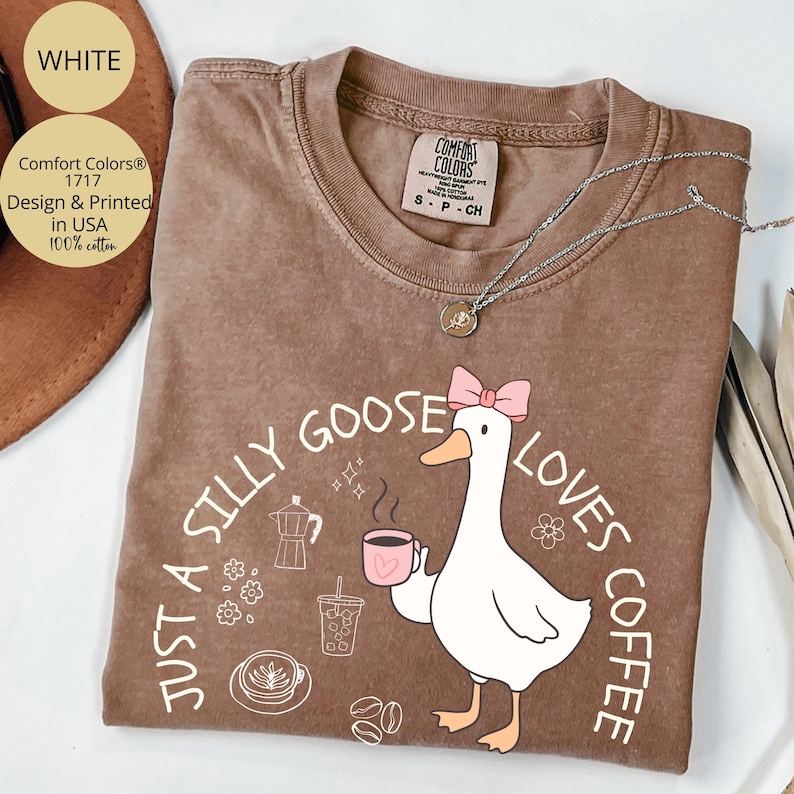 Puede incluir: Camiseta marr&oacute;n Comfort Colors con un dibujo de un ganso blanco con un lazo rosa, sosteniendo una taza de caf&eacute; rosa. El texto dice "Just a Silly Goose Loves Coffee". Un collar plateado est&aacute; sobre la camiseta.