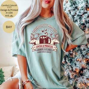 Puede incluir: Una camiseta verde claro con un dise&ntilde;o gr&aacute;fico de estilo vintage que presenta una taza de chocolate caliente, renos y el texto "North Pole IVF Mom Club", "Cocoa & Miracles", "Growing Hope Daily" y "Making Families Complete".