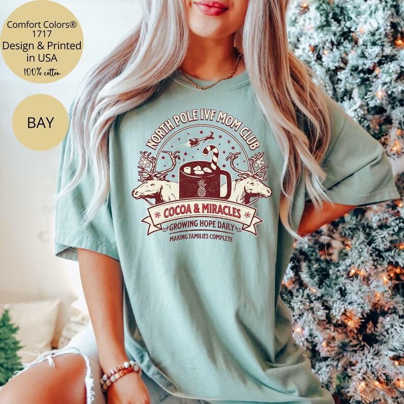 Puede incluir: Una camiseta verde claro con un dise&ntilde;o gr&aacute;fico de estilo vintage que presenta una taza de chocolate caliente, renos y el texto "North Pole IVF Mom Club", "Cocoa & Miracles", "Growing Hope Daily" y "Making Families Complete".