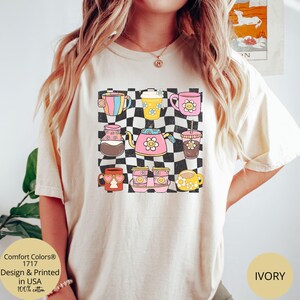 May include: A white t-shirt featuring a graphic of colourful mugs and teapots on a chequered background. The text "Comfort Colors&reg; 1717" is printed in the bottom left corner of the shirt. The text "IVORY" is printed in the bottom right corner of the shirt.
