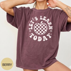 May include: Berry-colored t-shirt with the text "LET'S LEARN TODAY" in white. The design features a checkered apple in the center. The shirt has short sleeves and a relaxed fit.