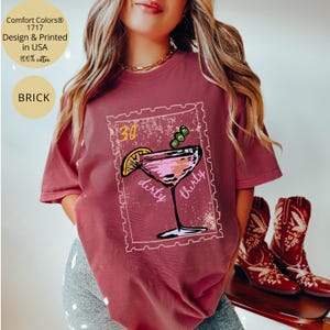 May include: Brick-colored t-shirt with a graphic of a cocktail, a lemon slice, and olives. The text "30 dirty thirty" is printed on the graphic. The shirt is made of 100% cotton and is designed and printed in the USA.