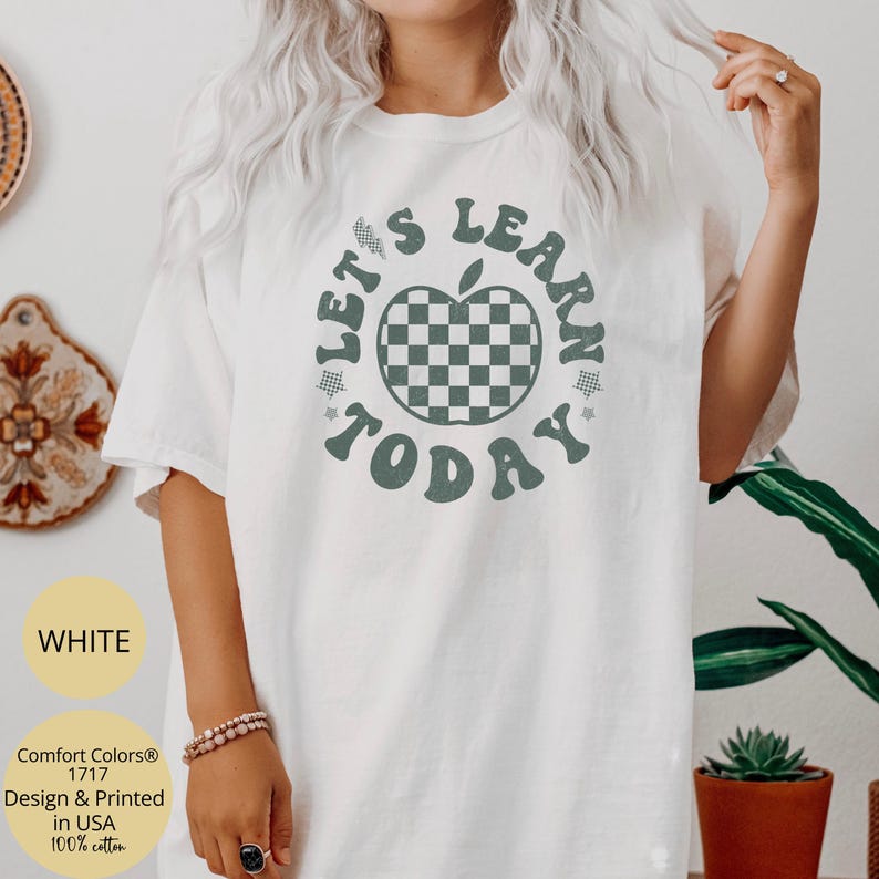 May include: White t-shirt with a green and white checkered apple graphic and the text "LET'S LEARN TODAY". The shirt is made of 100% cotton and is designed and printed in the USA.