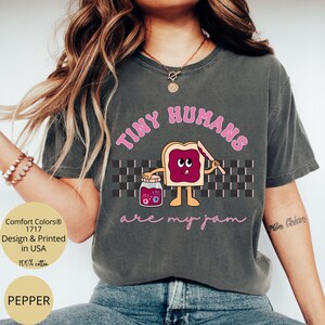 Pu&ograve; includere: T-shirt Comfort Colors grigio scuro con la scritta rosa "TINY HUMANS are my jam". La grafica presenta un toast dei cartoni animati con marmellata, un barattolo di marmellata e un coltello. La maglietta &egrave; 100% cotone e stampata negli Stati Uniti.