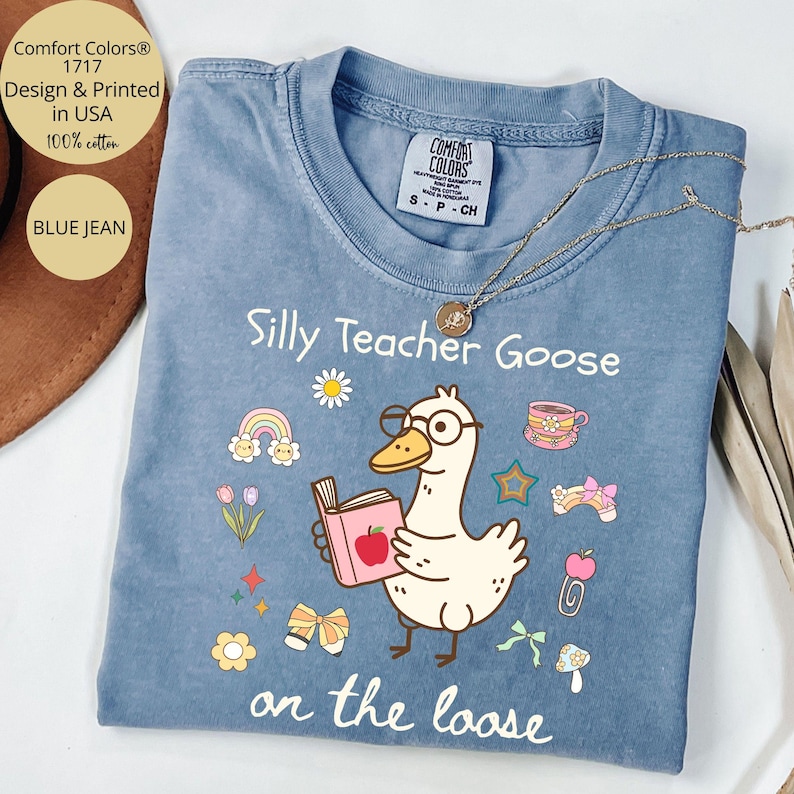 Pode incluir: Uma camiseta azul jeans com um ganso de desenho animado usando &oacute;culos e segurando um livro. O texto "Silly Teacher Goose on the loose" est&aacute; impresso na camiseta.