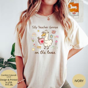 Pode incluir: Uma camiseta branca com um desenho animado de um ganso usando &oacute;culos e segurando um livro. O texto "Silly Teacher Goose on the loose" est&aacute; impresso na camiseta.