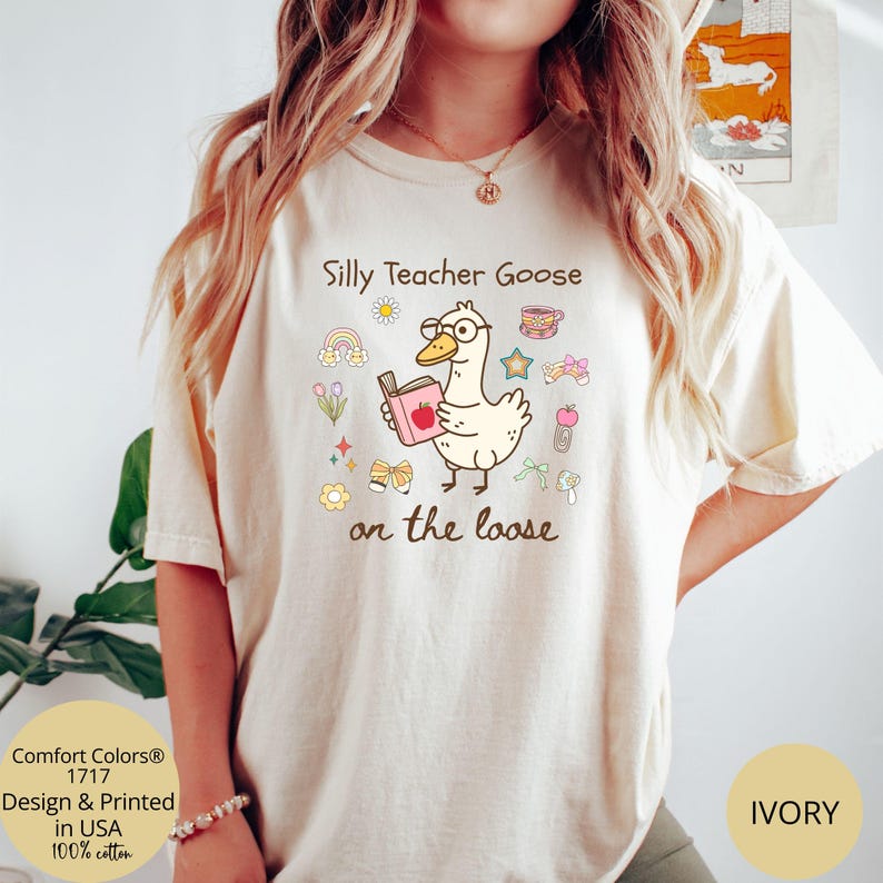 Pode incluir: Uma camiseta branca com um desenho animado de um ganso usando &oacute;culos e segurando um livro. O texto "Silly Teacher Goose on the loose" est&aacute; impresso na camiseta.