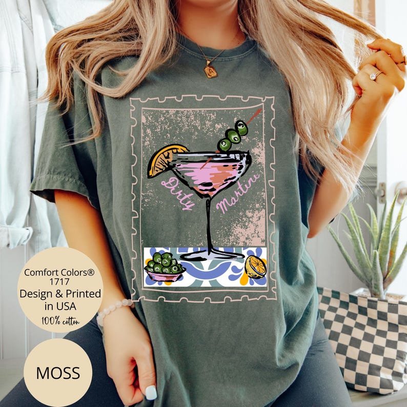 K&ouml;nnte beinhalten: Moosgr&uuml;nes T-Shirt mit einer Grafik eines rosa Dirty Martini Cocktails, Oliven und einer Zitronenscheibe. Das Design ist in einem Briefmarkenrahmen eingefasst. Das Shirt besteht aus 100% Baumwolle.