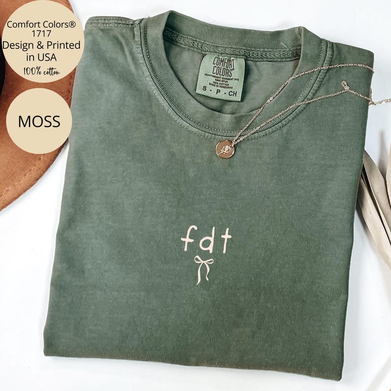 Peut inclure: Un t-shirt vert mousse Comfort Colors 1717 avec les lettres brod&eacute;es "fdt" et un n&oelig;ud. Un collier en argent avec un pendentif repose sur le t-shirt. Le t-shirt porte une &eacute;tiquette "Comfort Colors" et "100% coton".