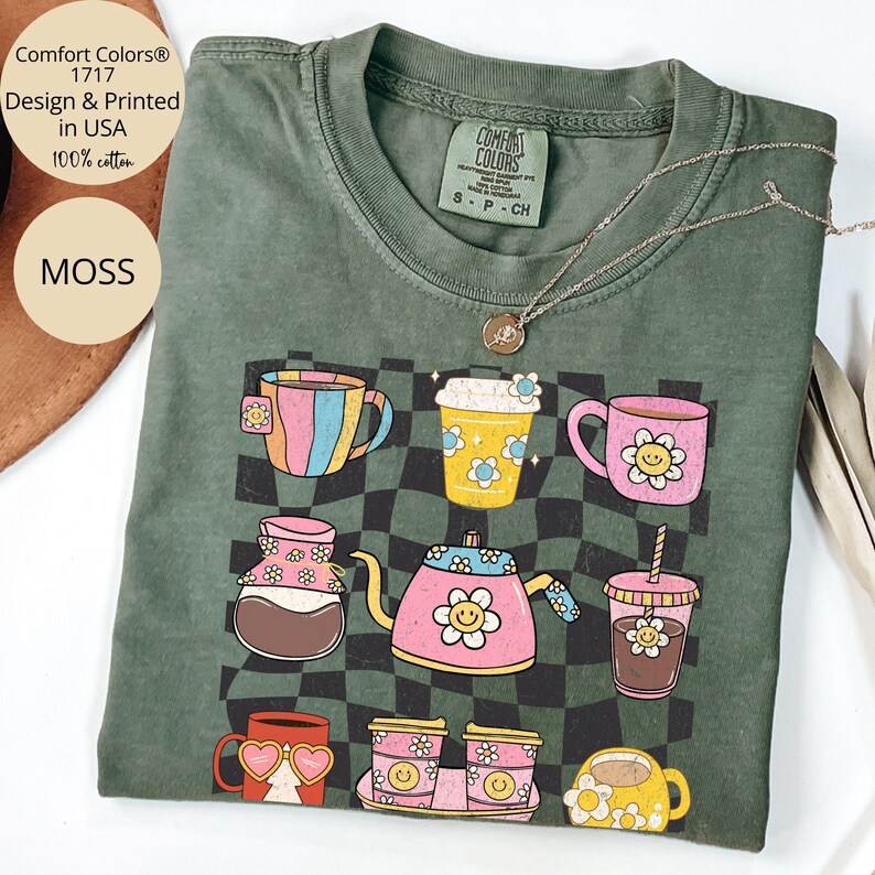 May include: A green t-shirt with a chequered pattern and a design of various coffee mugs, teapots, and coffee cups. The design features a smiley face on each mug and teapot. The text "Comfort Colors&reg; 1717 Design & Printed in USA 100% cotton MOSS" is printed on the shirt.