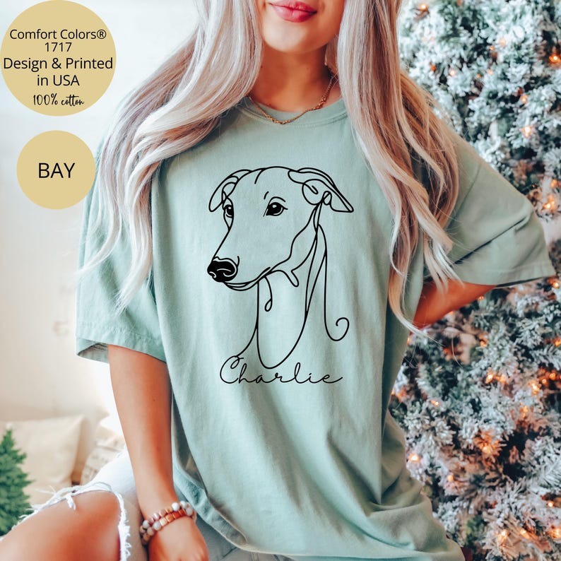 May include: A light green t-shirt featuring a minimalist black line drawing of a dog's head, with the name "Charlie" in script below. The shirt includes text that reads "Comfort Colors 1717 Design & Printed in USA 100% cotton" and the word "BAY".