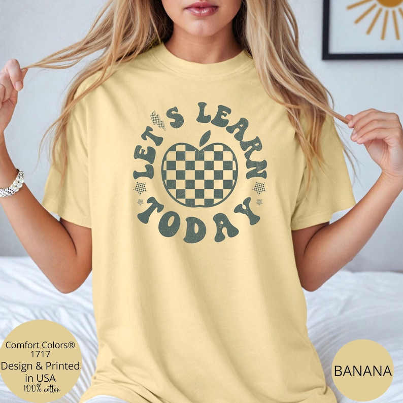 May include: A light yellow t-shirt with the text "LET'S LEARN TODAY" surrounding a checkered apple graphic. The shirt is made of 100% cotton and is printed in the USA. The shirt is labeled "BANANA".