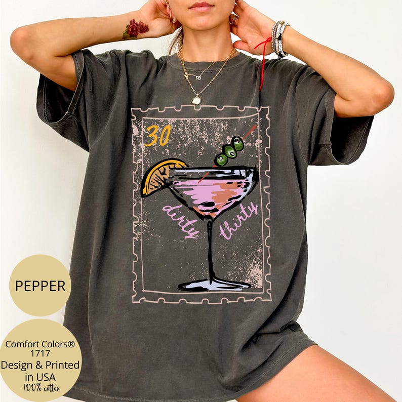 May include: A dark gray t-shirt with a cocktail graphic, featuring a pink drink with an olive garnish and a lemon slice. The text "30 dirty thirty" is displayed within a stamp-like border. The shirt is made of 100% cotton.