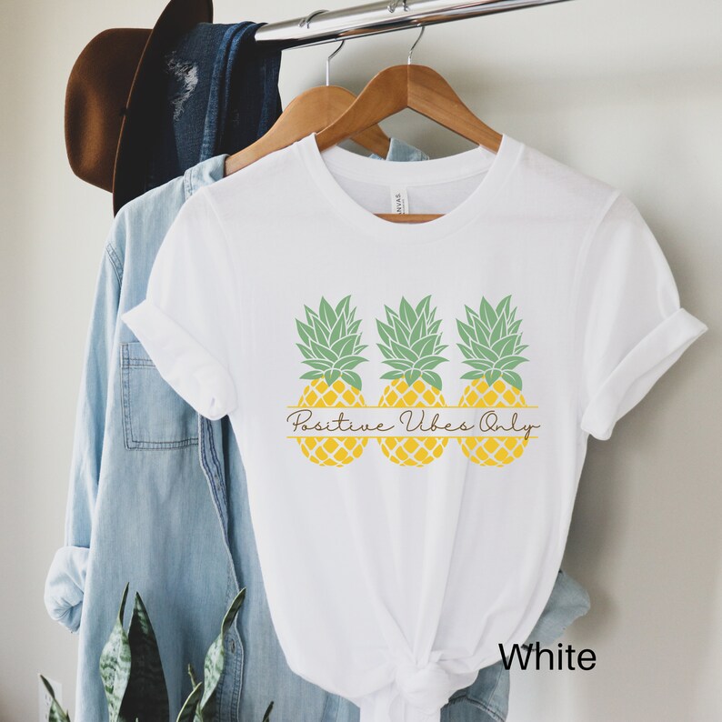 May include: White t-shirt featuring a design of three yellow pineapples with green tops and the text "Positive Vibes Only" in brown script.