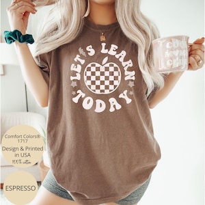 May include: Espresso-colored t-shirt with the text "LET'S LEARN TODAY" in a retro font. The design includes a checkered apple graphic. The shirt is made of 100% cotton and is designed and printed in the USA.