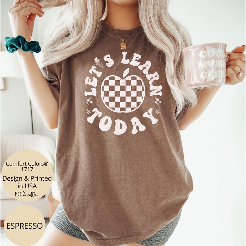 May include: Espresso-colored t-shirt with the text "LET'S LEARN TODAY" in a retro font. The design includes a checkered apple graphic. The shirt is made of 100% cotton and is designed and printed in the USA.