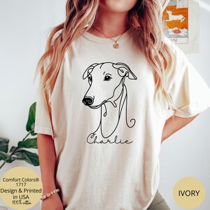 May include: Ivory-colored t-shirt featuring a minimalist black line drawing of a dog's head, with the name "Charlie" scripted below. The shirt includes text indicating it's designed and printed in the USA, made of 100% cotton, and the color is ivory.